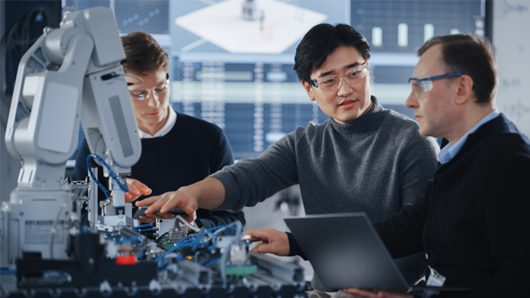 Three engineers in safety glasses collaborate on operating an advanced robotic system in a modern manufacturing lab, with one holding a laptop and others adjusting equipment.