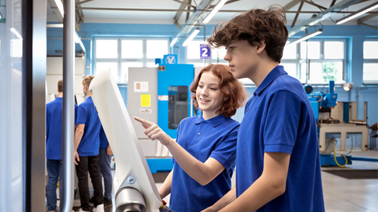 Two students in blue polos working together at a CNC machine interface in a modern manufacturing lab.