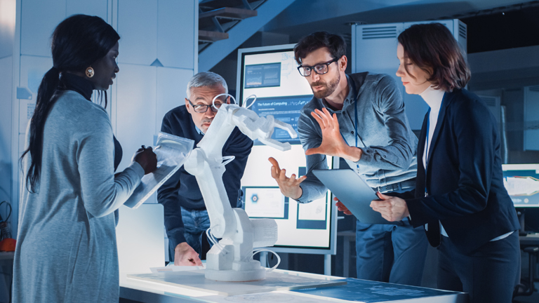 A small team reviews a robotic arm prototype in a modern lab, discussing training and workforce solutions supported by Tooling U-SME.