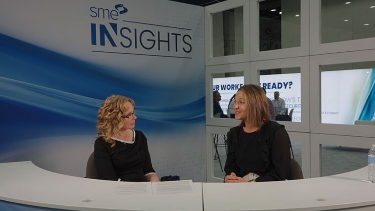 Becca O’Toole from Cengage sits behind a white desk and interviews with SME at the SME Insights booth